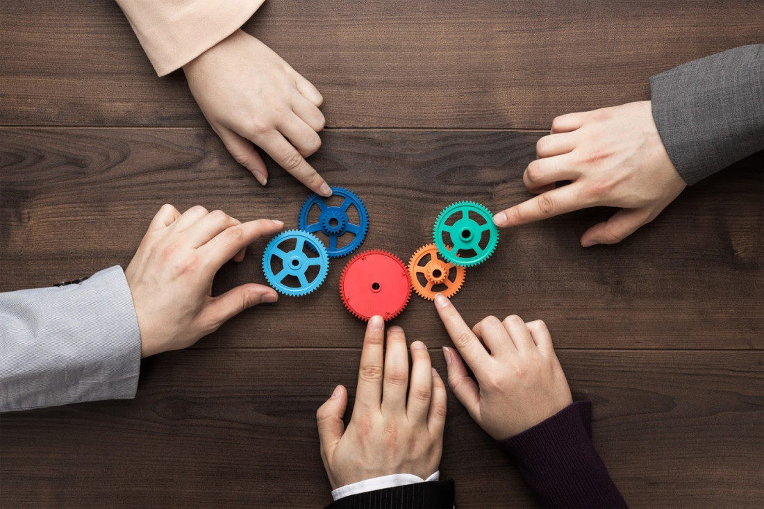 Teamwork-Konzept. Verschiedene Hände von Männern und Frauen verbinden bunte Zahnräder mit einem Arbeitsmechanismus auf braunem Holztisch-Hintergrund. 